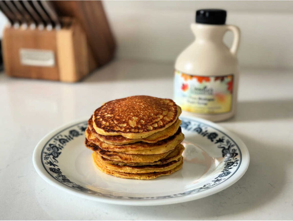 Pancakes stacked on a white plate with blue trip, maple syrup and knives are I the background, everything is sitting on a white countertop