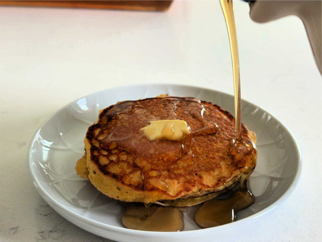 pancakes with syrup being poured on them and butter on a white plate on a white countertop